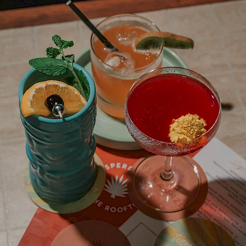 A trio of colorful cocktails on a table: a turquoise mug with lime and mint, an orange drink with a black straw, and a pink coupe with a yellow garnish, all atop coasters.