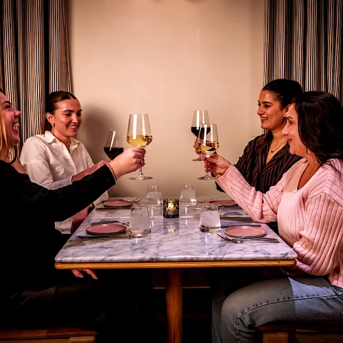A group of five friends sits around a table, toasting with wine glasses in a cozy dinner setting.