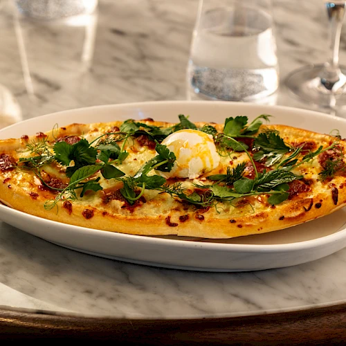 A baked boat-shaped dish (pizza or casserole) with melted cheese, herbs, and a dollop of creamy topping, served on a white plate on a marble table.