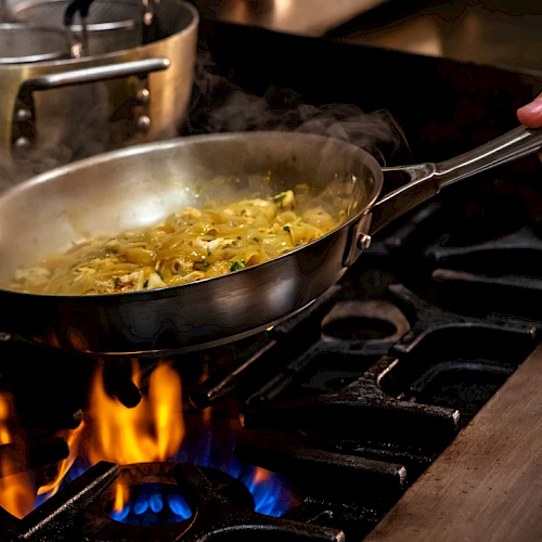 The image shows a person cooking in a frying pan over a gas stove, with flames under the pan and steam rising from the saut&eacute;ing ingredients, while a hand holds the pan handle, keeping it steady.