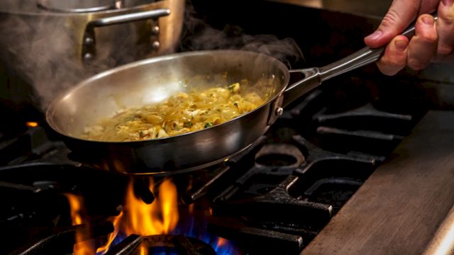 The image shows a person cooking in a frying pan over a gas stove, with flames under the pan and steam rising from the saut&eacute;ing ingredients, while a hand holds the pan handle, keeping it steady.