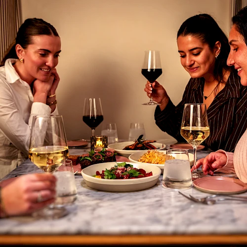 A group of five women enjoying a cozy dinner around a table with wine glasses, a plate of salad, and warm ambient lighting.