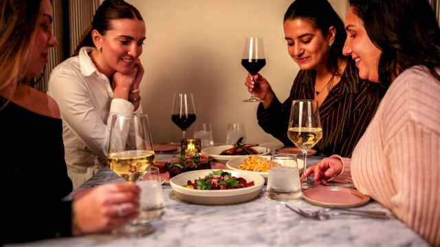 A group of five women enjoying a cozy dinner around a table with wine glasses, a plate of salad, and warm ambient lighting.