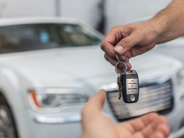 A person hands over car keys to another, with a car in the background, suggesting a vehicle handoff or ownership transfer.
