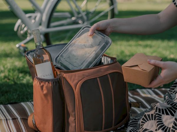A person packs a brown picnic bag with a meal container, a card, and a small box near a bicycle on a striped blanket at the park.