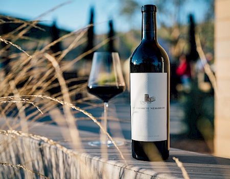 A dark wine bottle with a glass of red sits on a rustic board near a vineyard fence, blurred vines in the background.