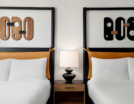 A modern hotel room with two beds, a bedside lamp, and decorative wall letters in black and wood tones.