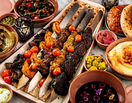 A platter of grilled meats and skewers with roasted chicken, pita, dips, and veggies like hummus and falafel.