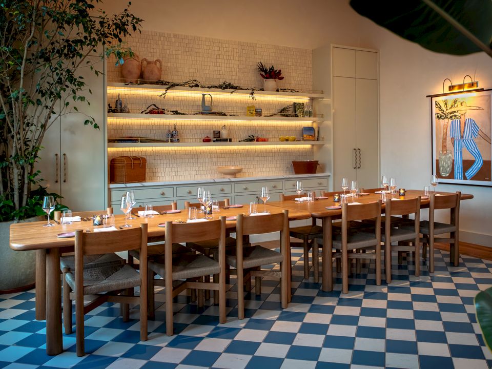 A bright restaurant dining area with a long wooden table, chairs, a blue-and-white tiled floor, and open shelves behind.