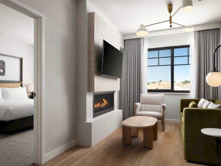 Modern hotel living room with couch, TV, fireplace, tall windows, curtains, and pendant lights.