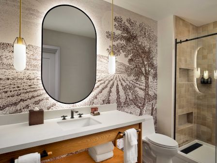 A modern bathroom with a white vanity and round mirror, a tree-lined wall mural, and a glass shower with tiled walls.