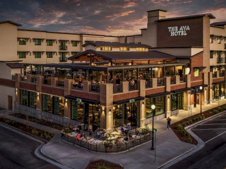 A hotel exterior at sunset with a &ldquo;THE AVA HOTEL&rdquo; sign, balconies, glass doors, and people dining on the patio.