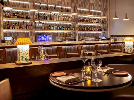 A stylish restaurant bar with shelves of bottles, warm pendant lights, and a set table with wine glasses and napkins.