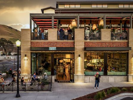 A modern restaurant patio at dusk with diners, a sign reading &ldquo;EMRE,&rdquo; and people seated by lit windows and a doorway.