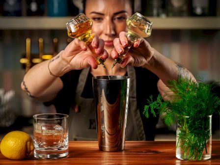 A bartender pours golden and green liquors into a metal cocktail shaker, with an herb sprig and lemon on the bar.