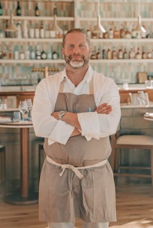 A bearded chef in a white shirt and gray apron stands with arms crossed in a warmly lit caf&eacute; with bookshelves behind him.