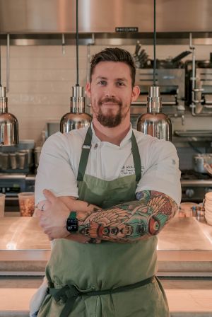 A bearded chef with sleeve tattoos poses in a kitchen bar with brewing equipment and glass bottles behind him.