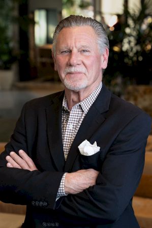 Portrait of an older man in a dark blazer and checkered shirt, arms crossed, with a white pocket square in a blurred office.
