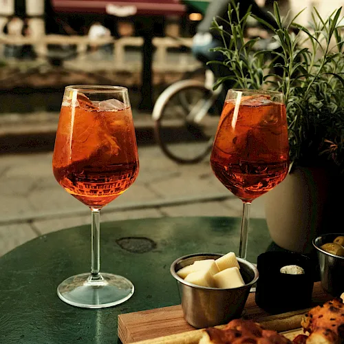 Two glasses of iced amber drink with rosemary sit by cheese and bread on a café table, ready for a toast.