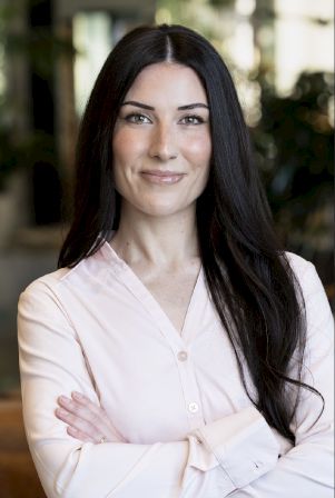 A smiling woman with long dark hair in a white blouse, arms crossed, against a blurred office-like background.