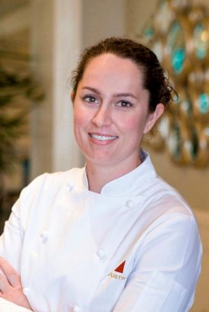 A smiling woman in a white chef jacket with &ldquo;JUSTIN&rdquo; on it, standing with arms crossed in a warmly lit background.
