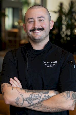 A smiling chef in a black hotel jacket, arms crossed to show tattoos, posed against a softly blurred interior background.