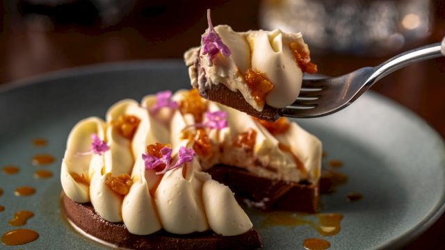 A fork holds a bite of chocolate cake topped with white cream, caramel drizzle, and edible purple flowers.