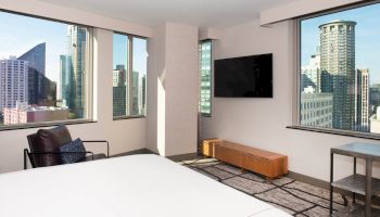 A modern hotel room with a large bed, TV, and armchair, featuring large windows with a cityscape view and contemporary decor.