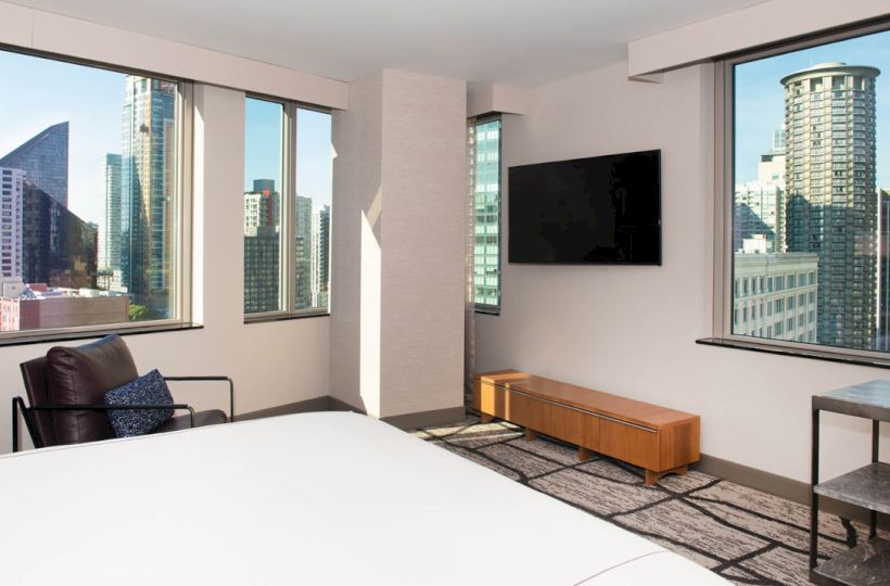 A modern hotel room with a large bed, TV, and armchair, featuring large windows with a cityscape view and contemporary decor.