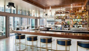 The image shows a modern bar with high stools, a mirrored back wall with bottles, and large windows offering a city view.