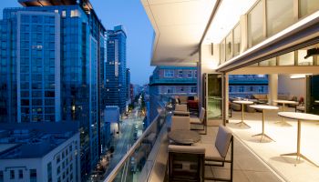 A modern cityscape at dusk with tall buildings and a sleek balcony featuring tables and chairs.