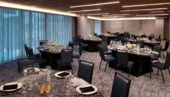 The image shows an elegantly arranged dining area with round tables, chairs, and neatly set tableware under dim lighting.