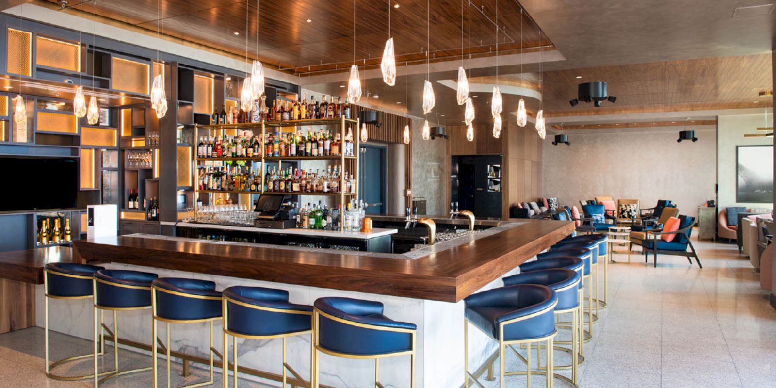 A modern bar with a wooden counter, blue bar stools, pendant lights, and a well-stocked bar backdrop, set in a spacious lounge area.