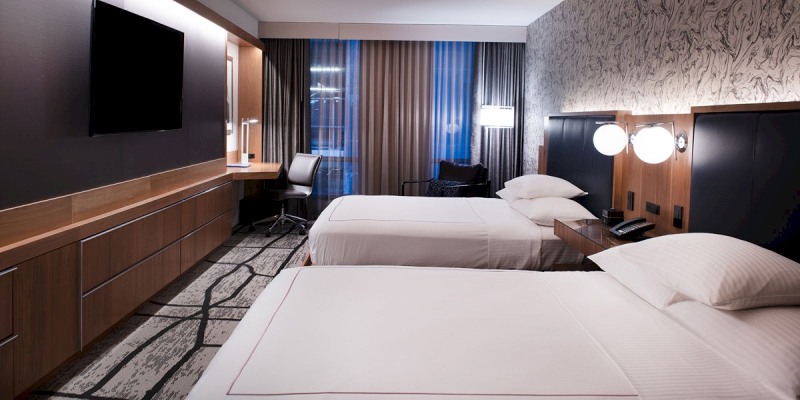 A modern hotel room featuring two beds, a desk with a chair, a flat-screen TV, lamps, and patterned decor on the walls and floors.