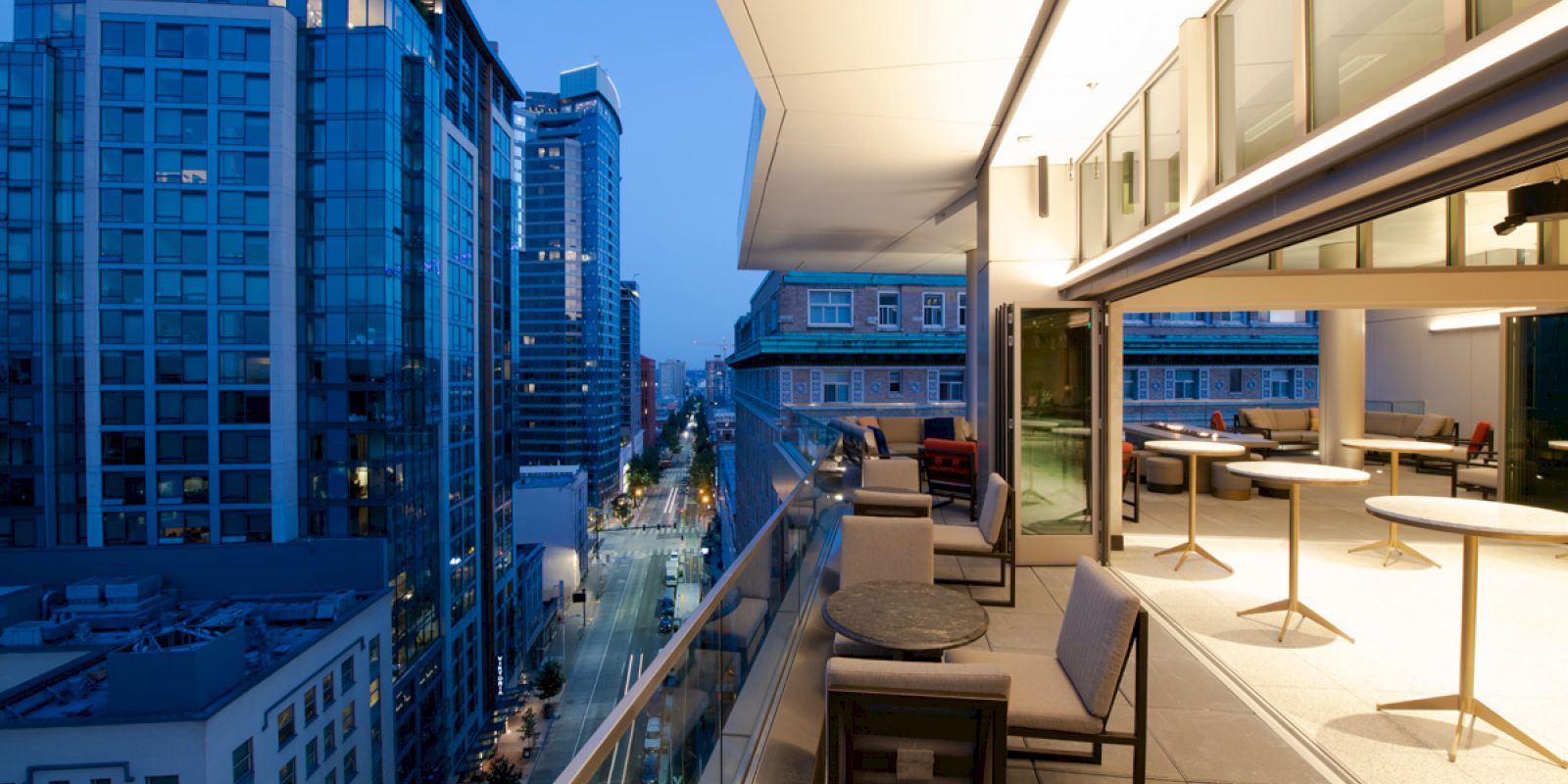 A modern cityscape at dusk with tall buildings and a stylish balcony overlooking a street, featuring seating areas and soft lighting.