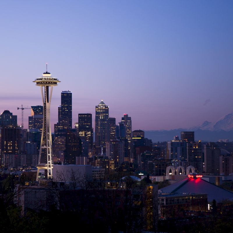 A cityscape during dusk with a prominent tower, tall buildings, and a mountain in the background under a clear sky.