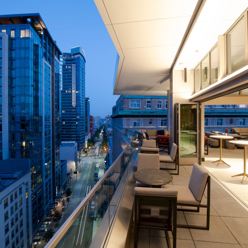 A modern cityscape view from a high balcony with elegant outdoor seating, overlooking a street lined with tall buildings at dusk.
