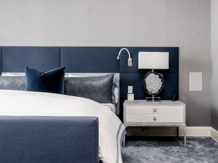 A modern bedroom with a navy bed, light gray nightstand, wall lamp, and decorative lamp on a carpeted floor.