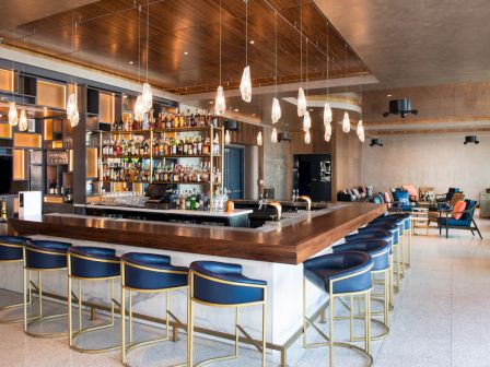 A modern bar with a central counter, blue stools, hanging lights, and a well-stocked liquor shelf, set in a spacious, stylish lounge.
