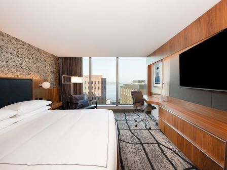 A modern hotel room features a bed, desk, chair, large TV, and city view through a window, with a stylish design and decor elements.