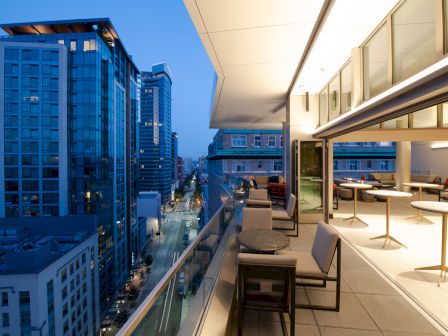 A modern urban balcony at dusk with seating overlooks a city street lined with tall buildings, under a blue sky.