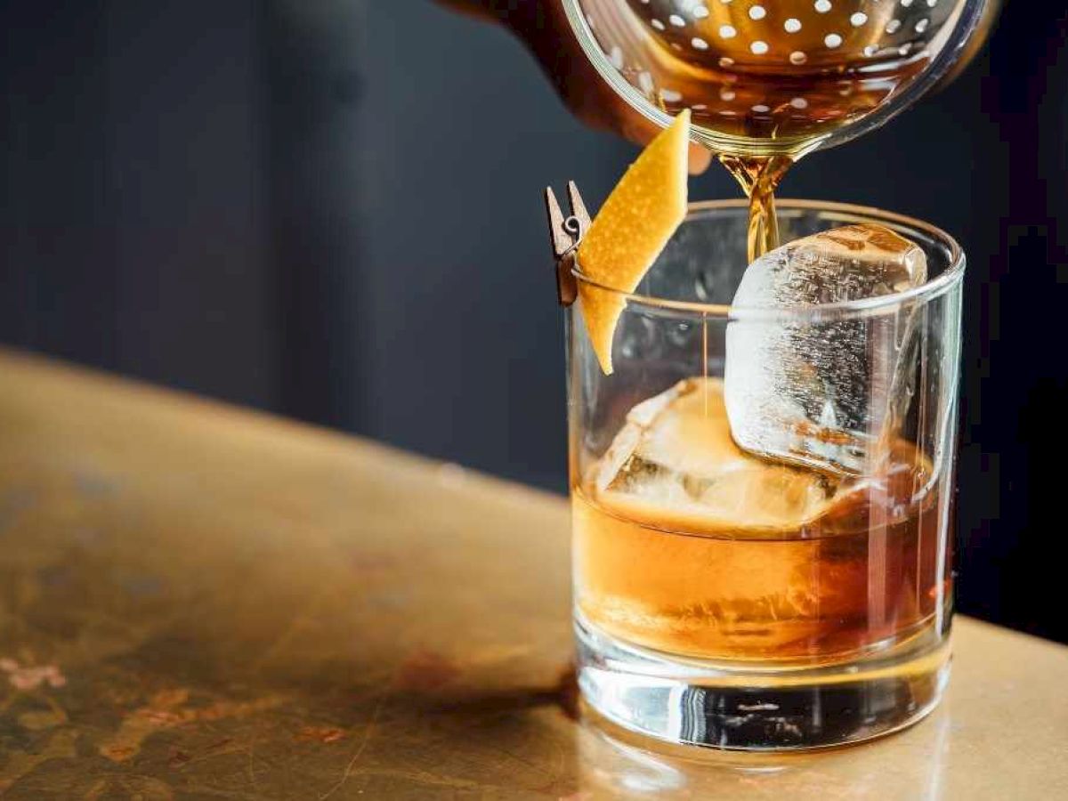 A cocktail being poured into a glass with ice and a citrus peel garnish, all on a wooden surface.