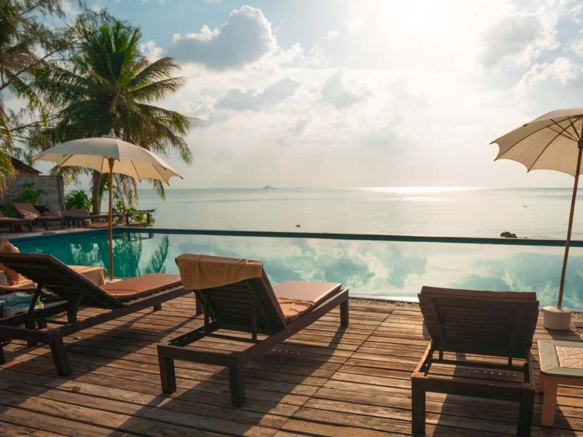 A serene seaside scene with lounge chairs, umbrellas, palm trees, and a pool overlooking a calm ocean under a bright sky.