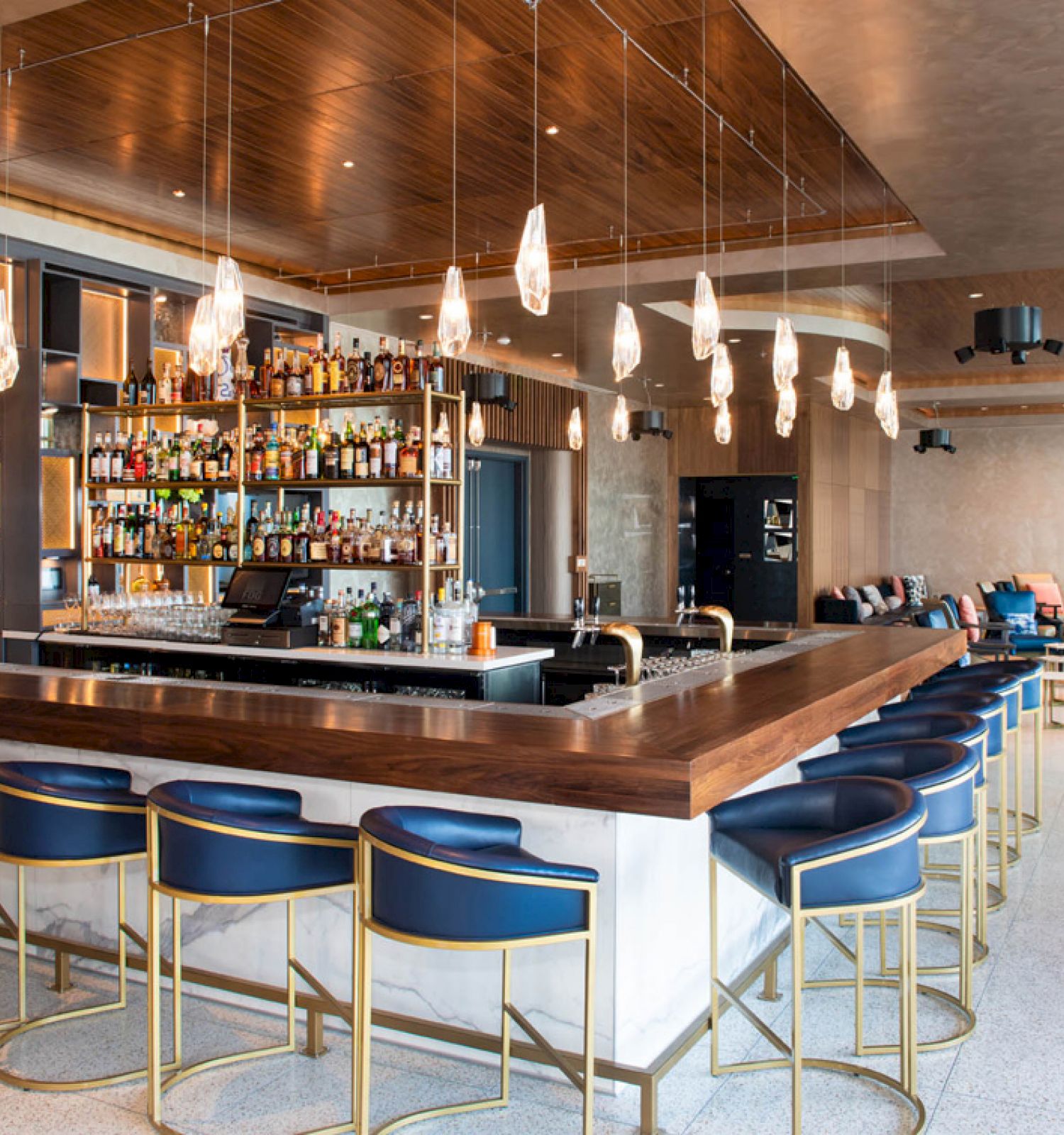 A modern bar with blue bar stools, pendant lights, a stocked shelf with bottles, and seating areas in a spacious, stylish interior.