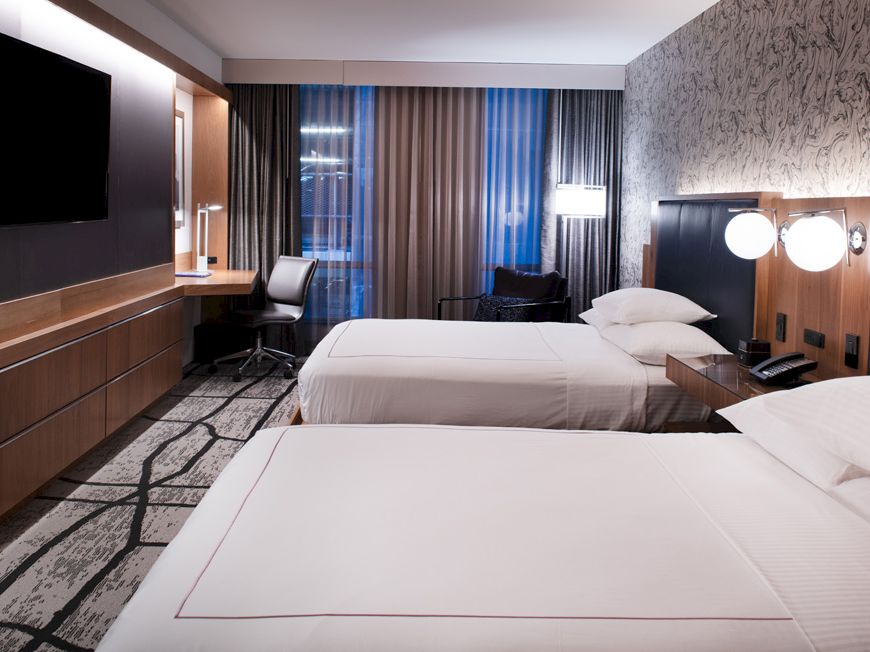 The image shows a modern hotel room with two beds, a TV, desk, and stylish decor, featuring a patterned carpet and wallpaper.