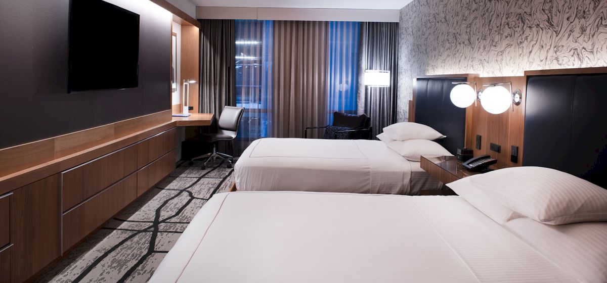 The image shows a modern hotel room with two beds, a TV, desk, and stylish decor, featuring a patterned carpet and wallpaper.