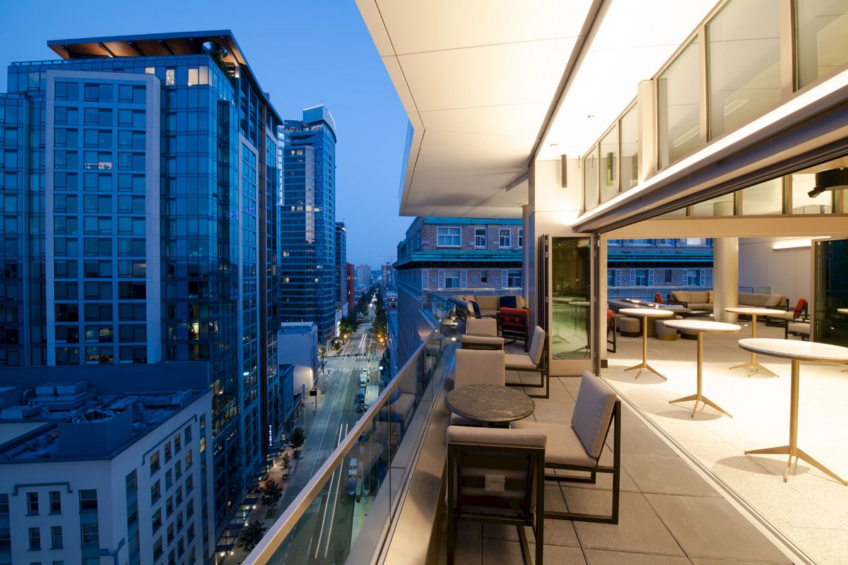 A cityscape view with tall buildings and a modern rooftop terrace featuring tables and chairs overlooking a street at dusk.