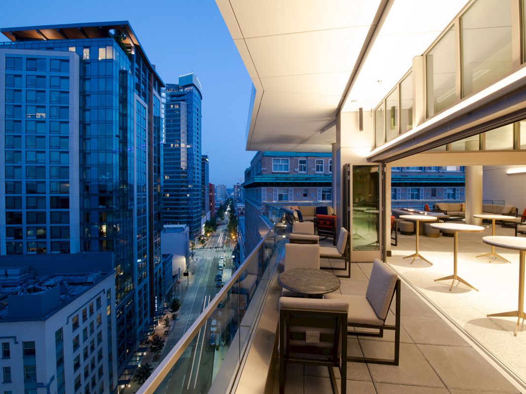 A cityscape view with tall buildings and a modern rooftop terrace featuring tables and chairs overlooking a street at dusk.