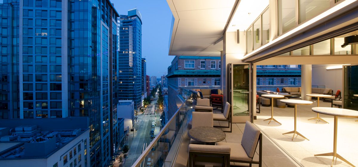 A cityscape view with tall buildings and a modern rooftop terrace featuring tables and chairs overlooking a street at dusk.