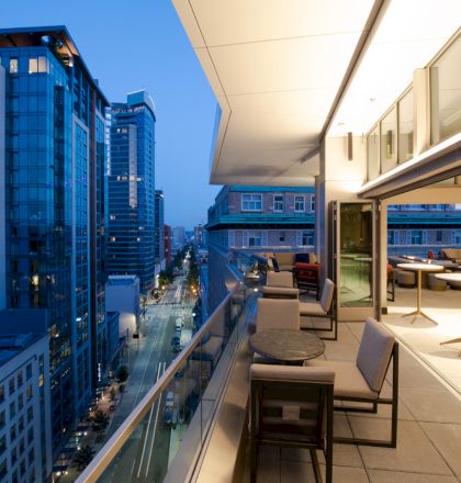 A modern urban balcony overlooking a city street at dusk, with outdoor seating and large windows opening into a lit interior space.